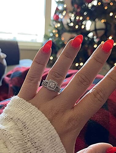 Hand with red nails wearing a diamond ring in front of a Christmas tree.